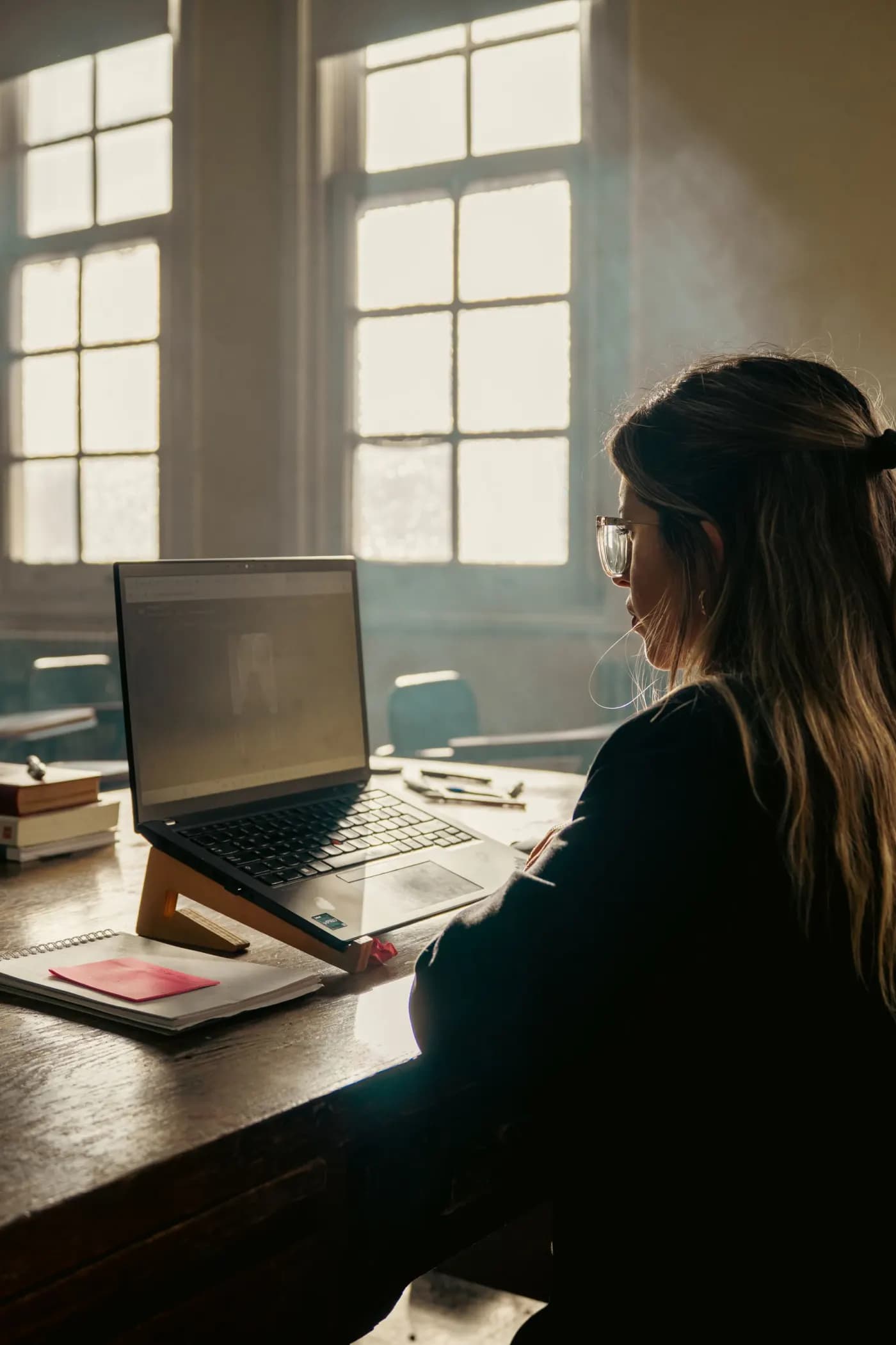 En elev set bagfra ved sin bærbare computer i et klasselokale, dagslys gennem høje vinduer, notesbog og bøger på bordet